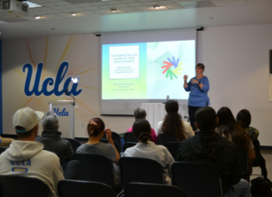 The image captures the lecturer, Donalda Ammons, signing in front of an audience. Donalda is standing on a stage, with a PowerPoint slideshow behind her that reads “Celebrating 100 years of the Deaflympics, Preserving Institutional Independence.” The slideshow also features the Deaflympics logo, which consists of hands forming a circle with blue, yellow, green, and red features.